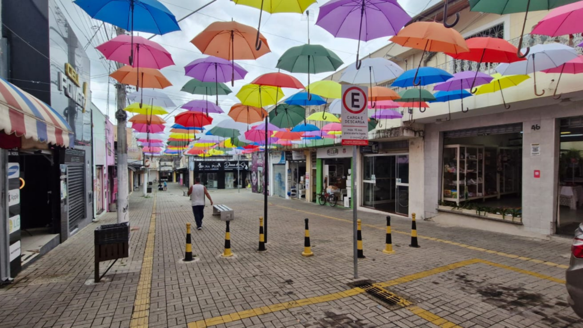 Imagem do Centro Comercial 10 de Julho em Pindamonhangaba, decorado com guarda-chuvas coloridos, que trazem um novo visual ao espaço e atraem visitantes e turistas. A iniciativa visa revitalizar o comércio local e promover um ambiente acolhedor.